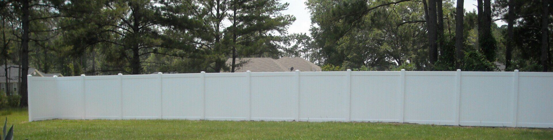 White fence in a grassy yard, with trees and the roof of a house in the background.