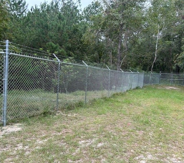 Chain-link fence topped with barbed wire, set in a grassy area with trees in the background.