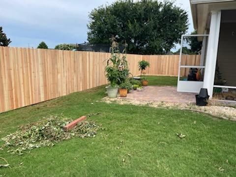 Wooden fence encloses a green lawn and brick patio with potted plants near a house under a cloudy sky.