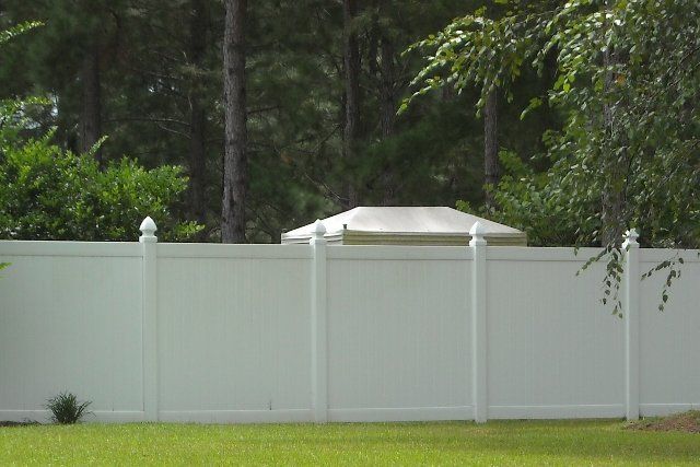 White vinyl fence in front of green grass and trees. A light-colored structure can be seen behind the fence.