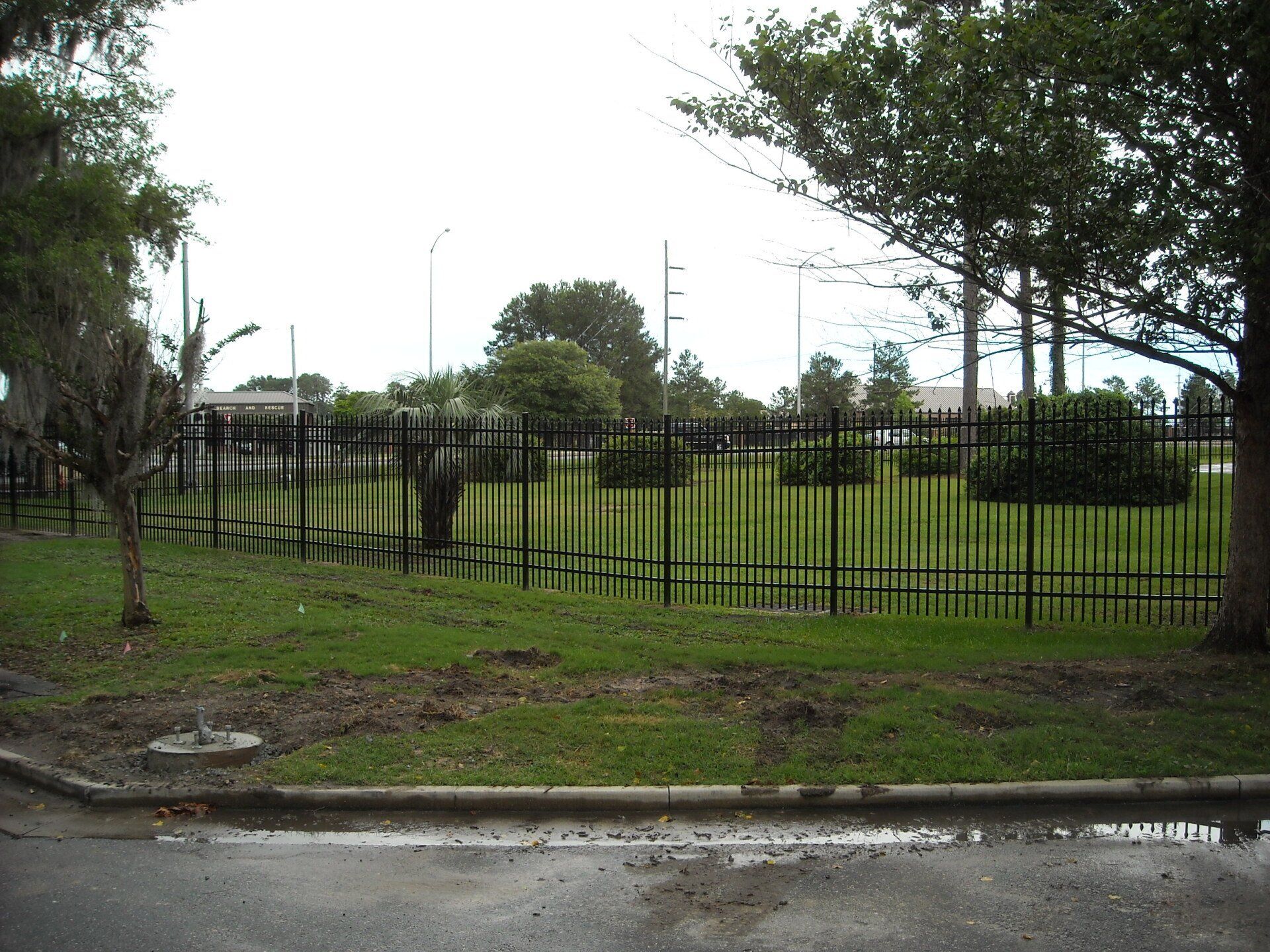 Black metal fence bordering a grassy area with trees, overcast sky, and a wet road in the foreground.