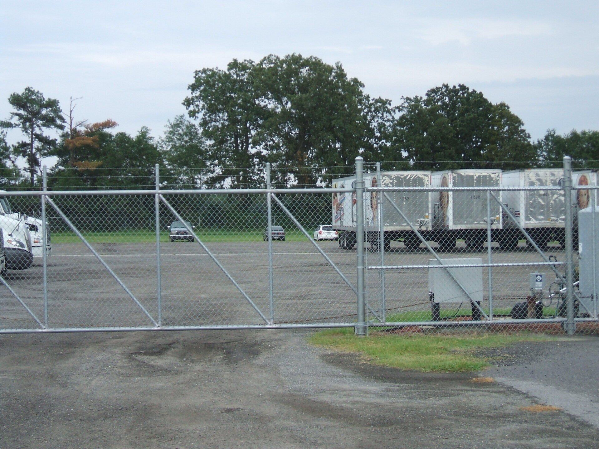 Chain-link fence with gate, enclosing an industrial yard. Semitrucks and trailers visible. Cloudy sky, grassy area.