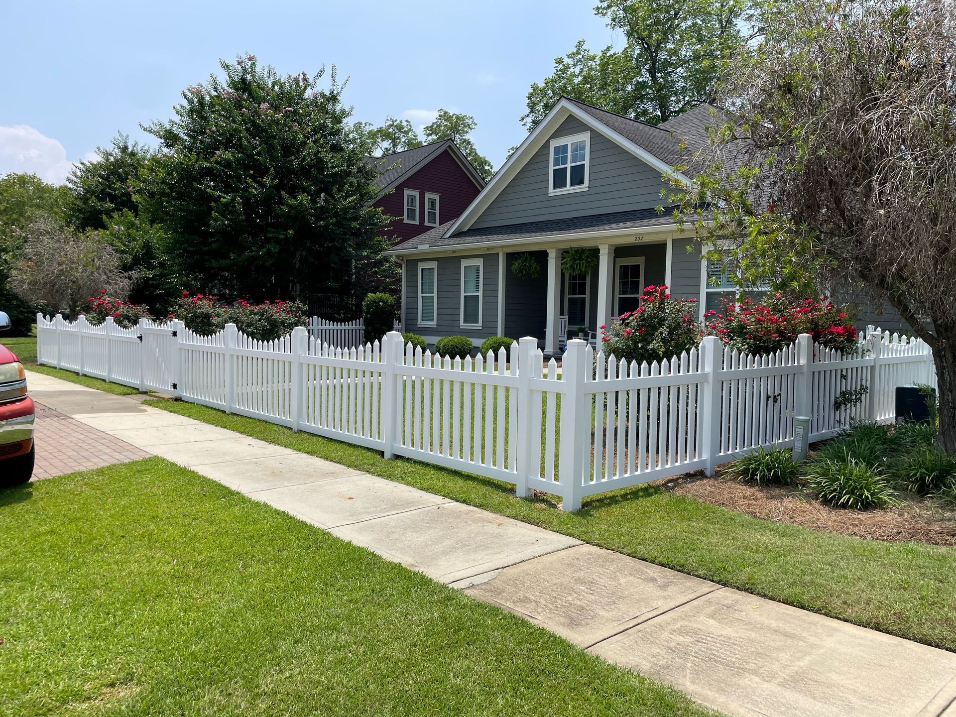 White picket fence surrounds a green house with hanging red flower baskets. Sidewalk and green grass visible.