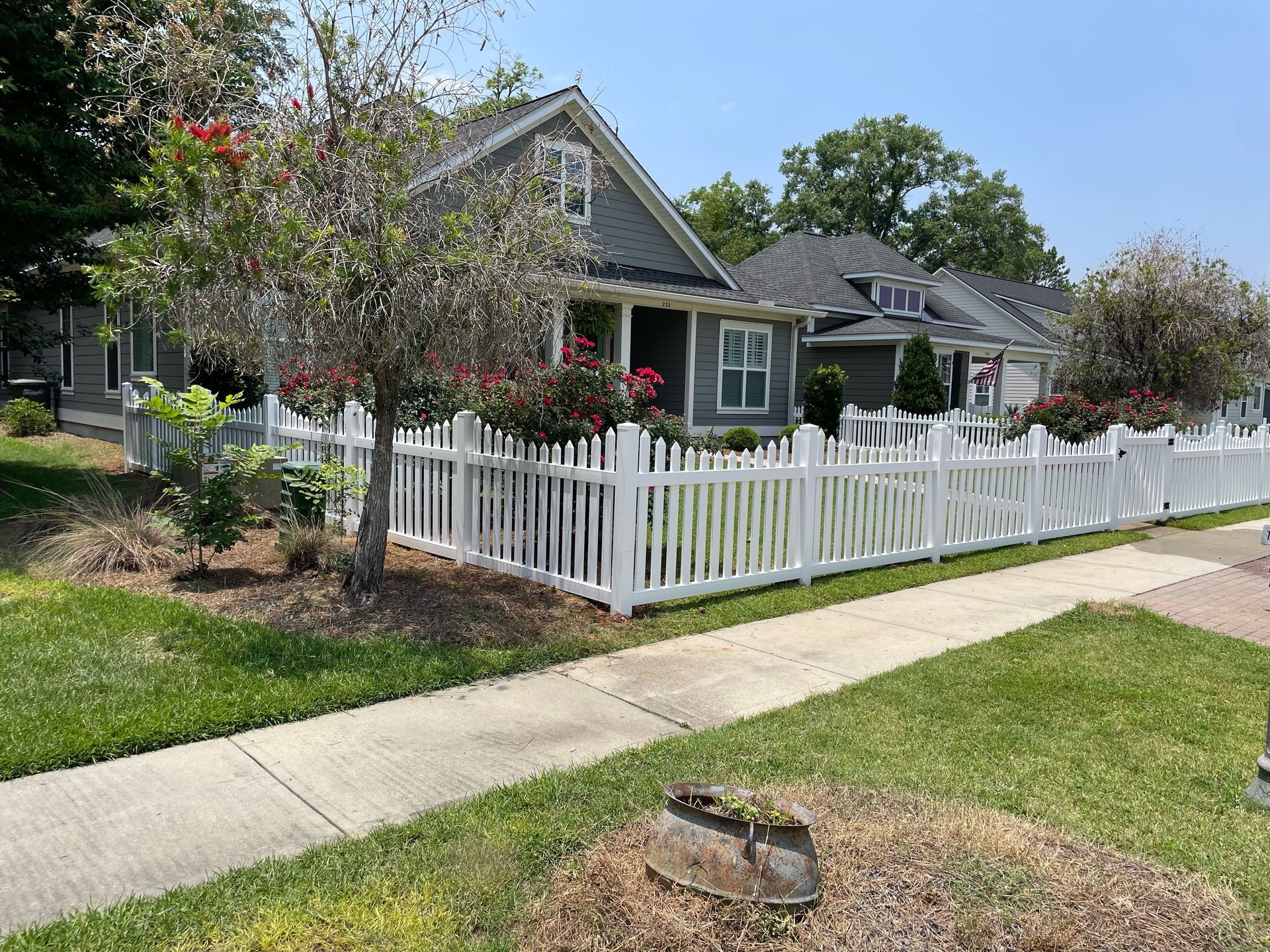 White picket fence in front of gray houses with red flowers and green grass on a sunny day.
