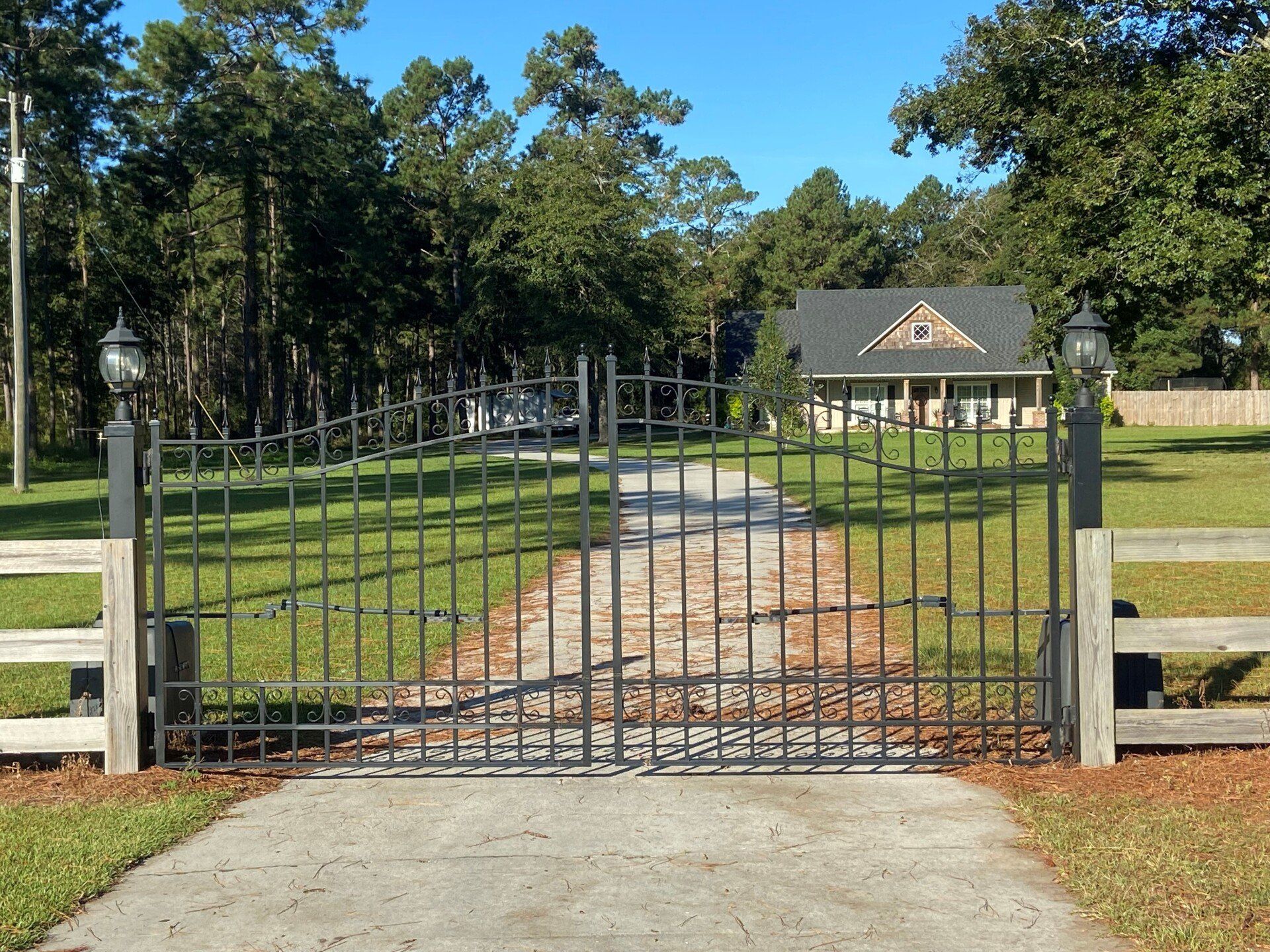 Black metal gate with lights, leading to a house down a concrete driveway, surrounded by a grassy yard and trees.