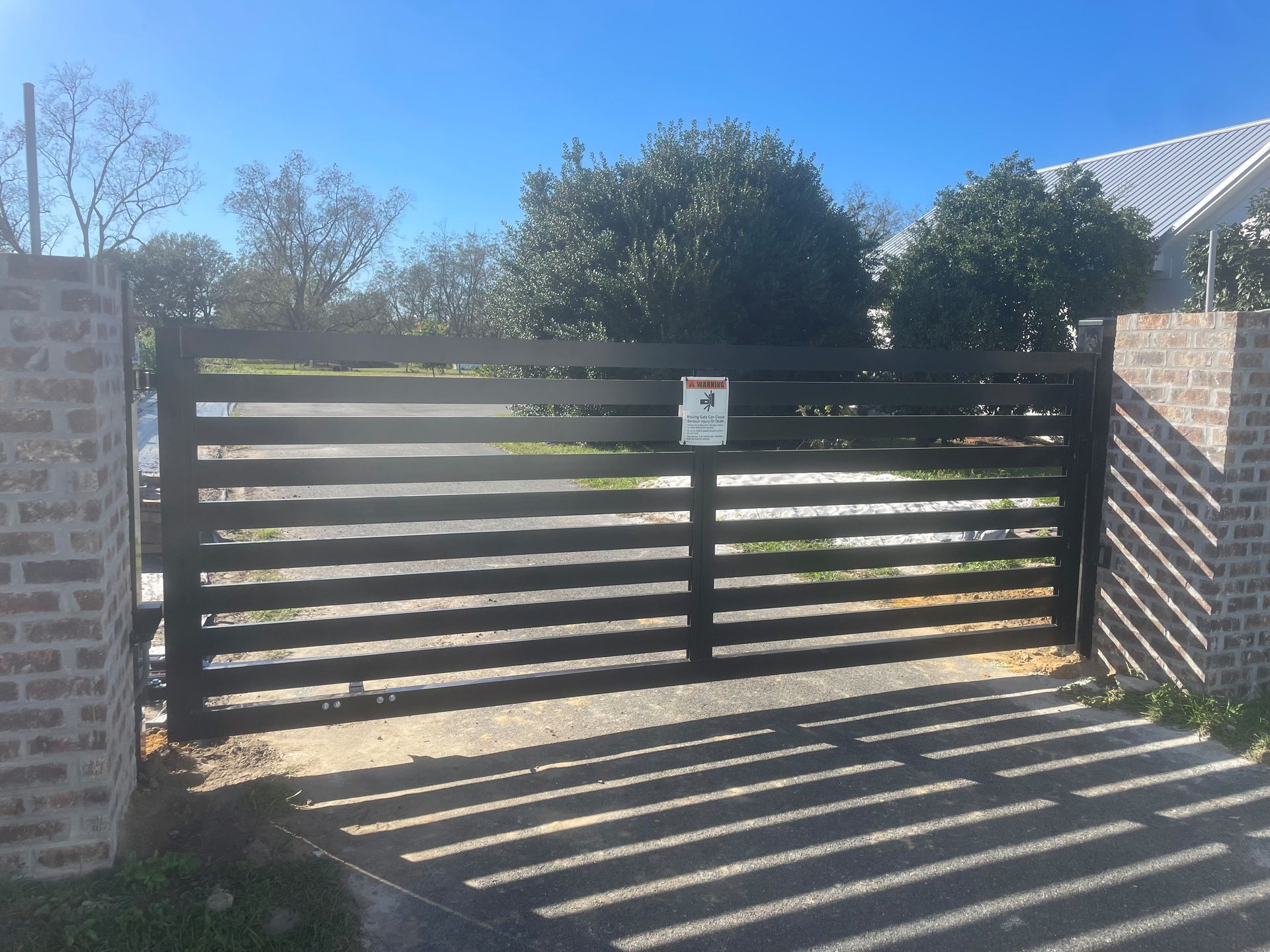 Black horizontal slat gate between brick pillars on a driveway, with a security sign.