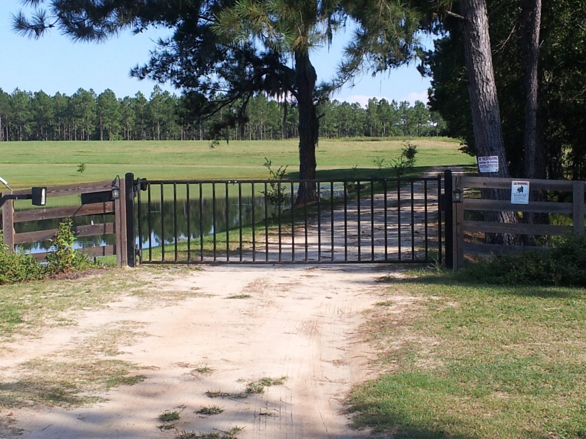 Black metal gate across a dirt driveway leading to a grassy field with trees.