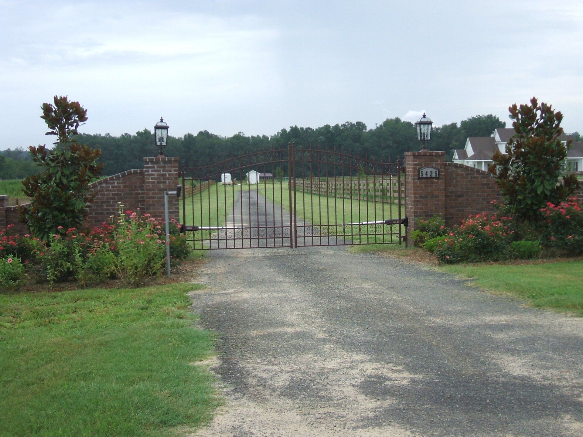 Gated entrance to a gravel driveway. Brick pillars with lights flank the metal gate, with flowers and trees.