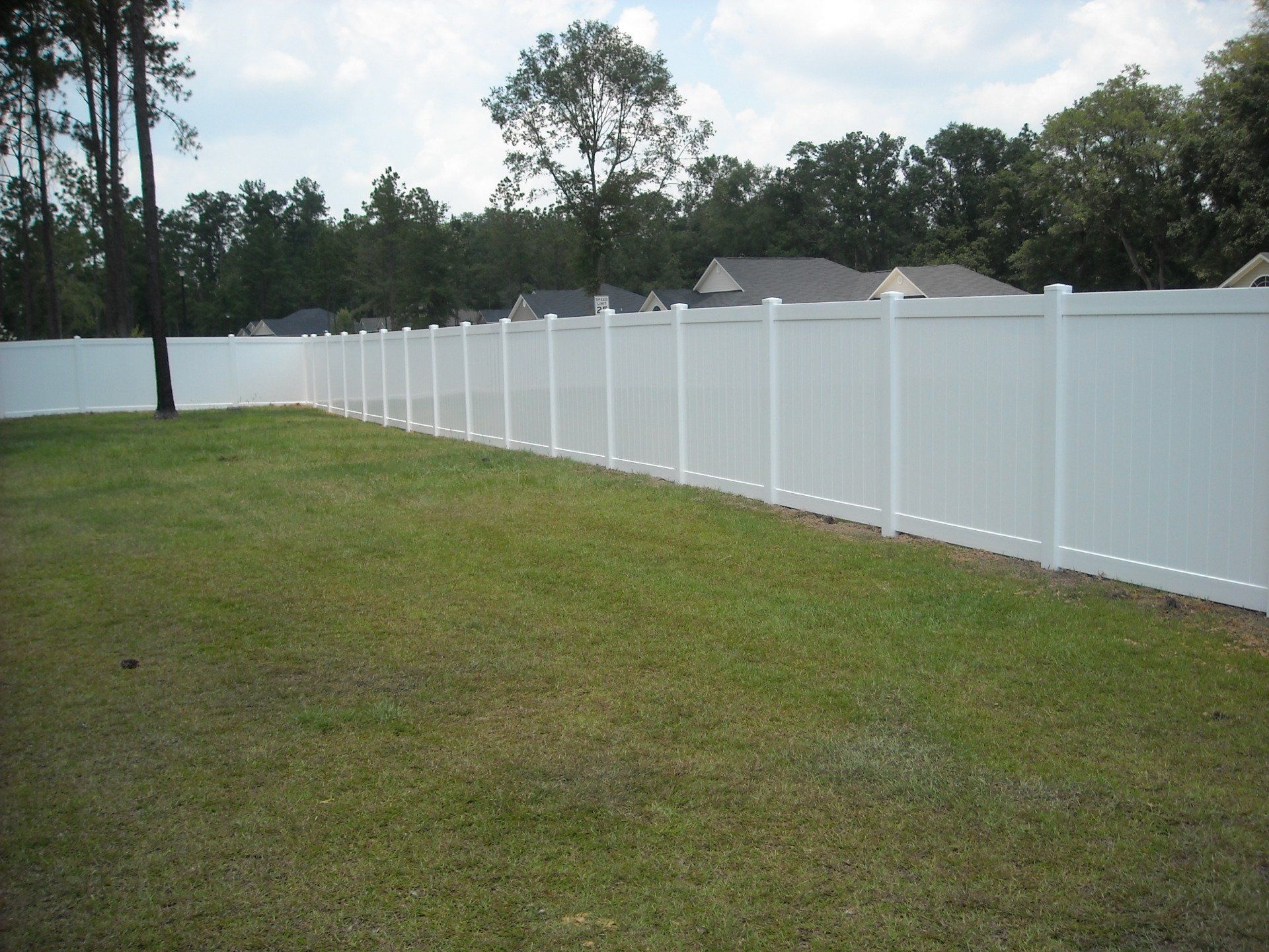 White vinyl fence surrounds a grassy backyard, trees and houses in the background.