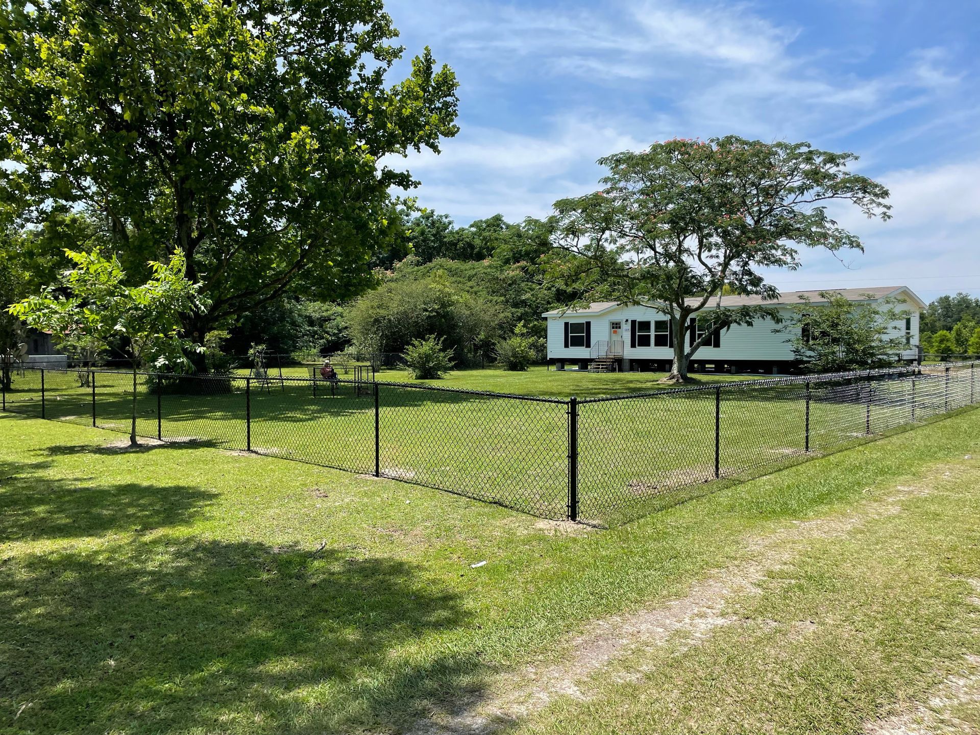 White manufactured home behind a black fenced-in grassy yard, with a large tree and greenery in the background.