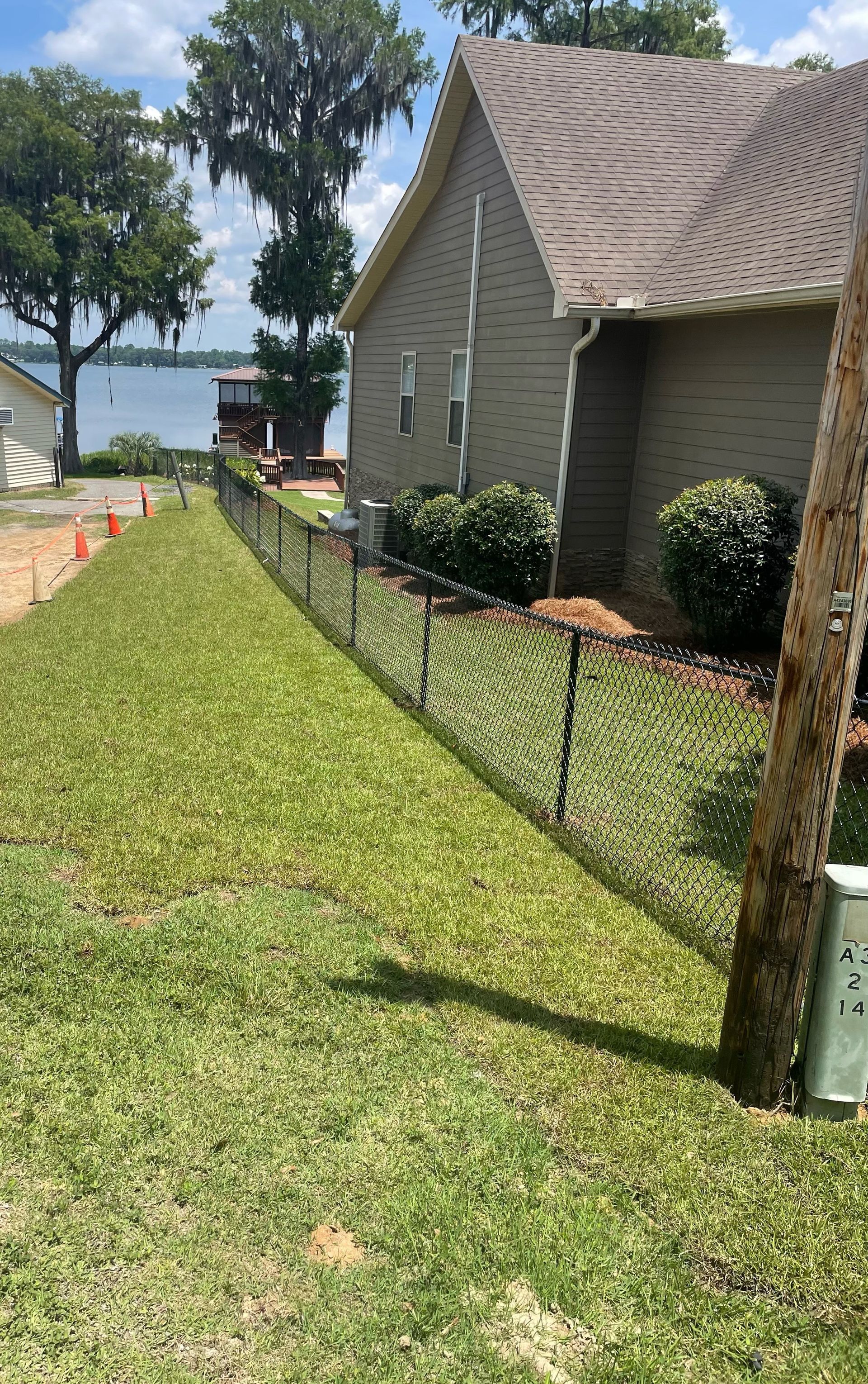 A black chain-link fence borders a grassy yard next to a beige house, with a lake in the background.