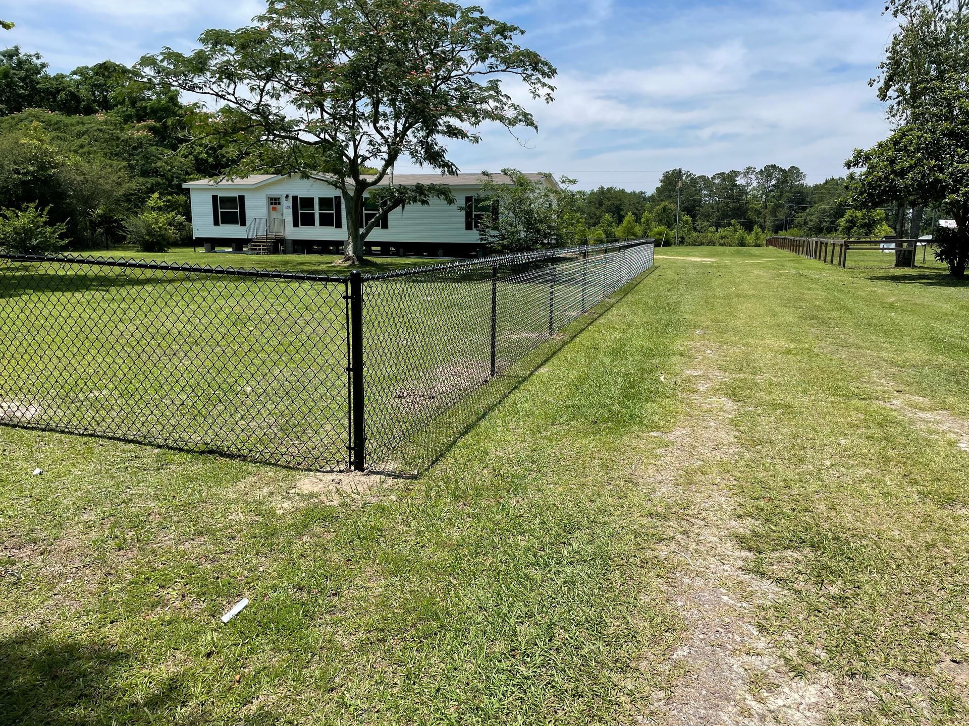 Black chain-link fence bordering a grassy area with a mobile home in the background. Sunny day.