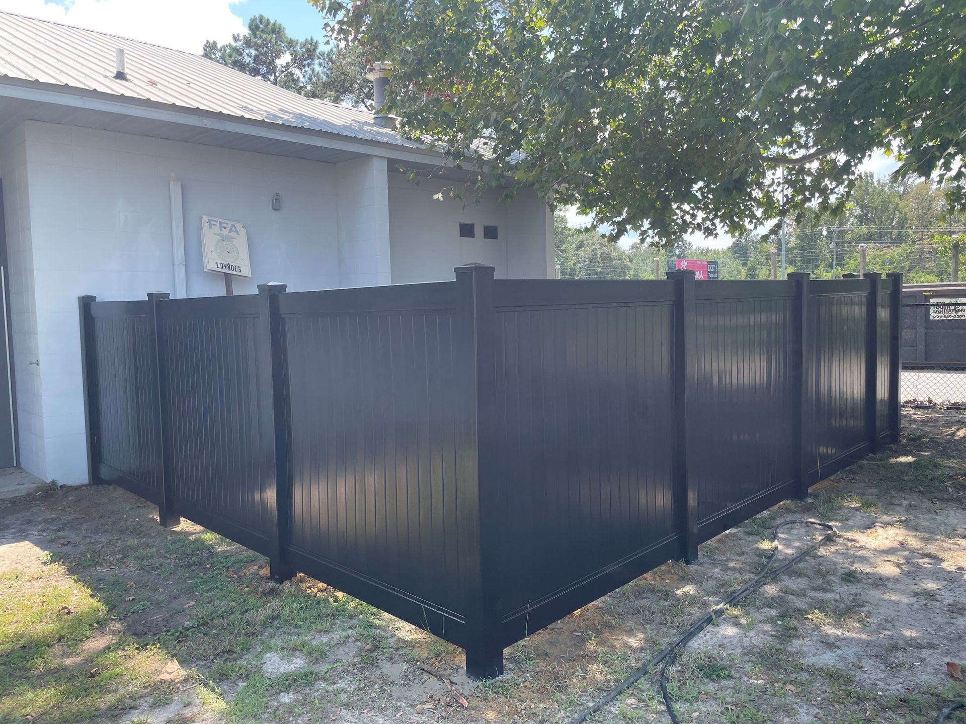 Black vinyl fence encloses a backyard with a gray building, grass, and a tree.