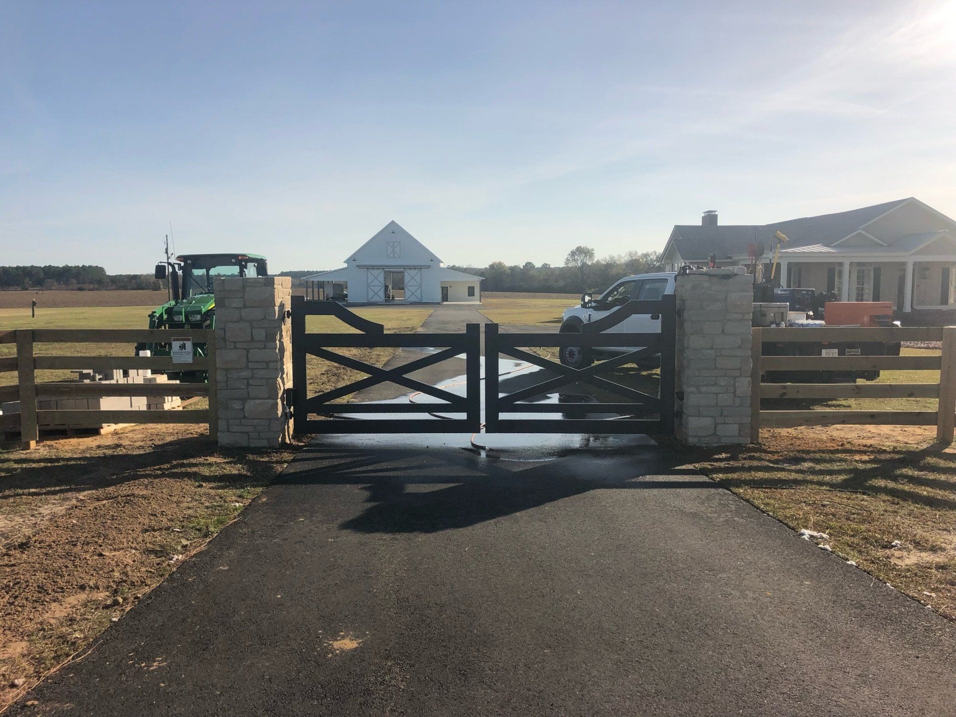Black wooden gate, stone columns, asphalt driveway leading to a field with a barn and a house.