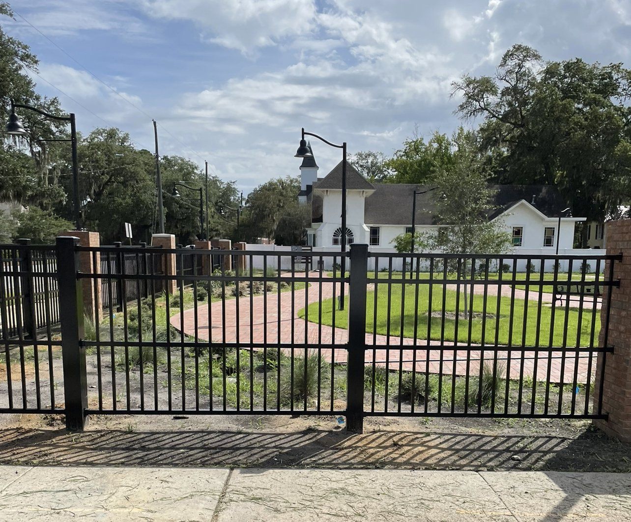Black fence in foreground with grassy area, brick pathway and white house in the background. Overcast sky.