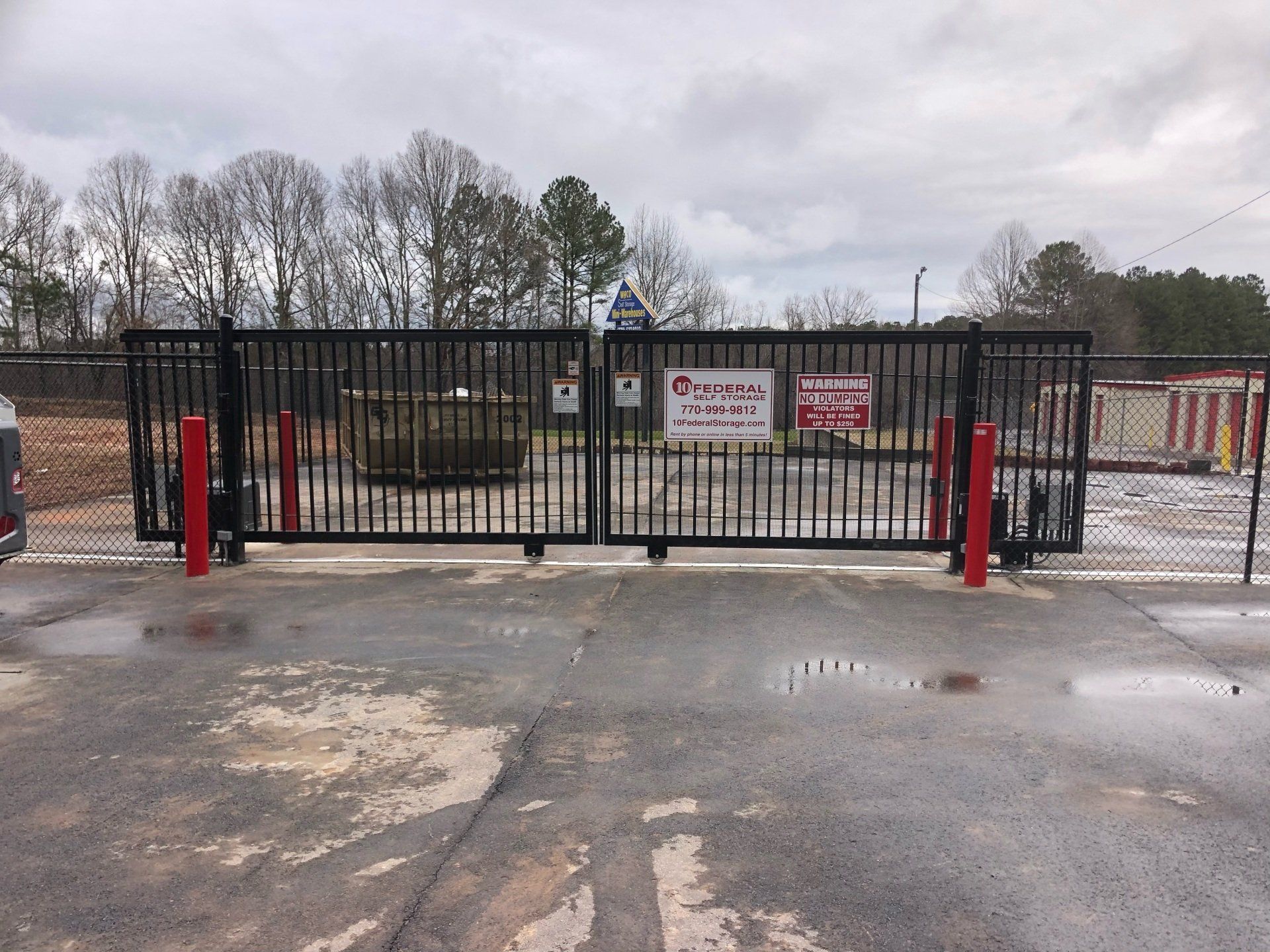 Black metal gate with red posts, leading to a storage facility with a cloudy sky.