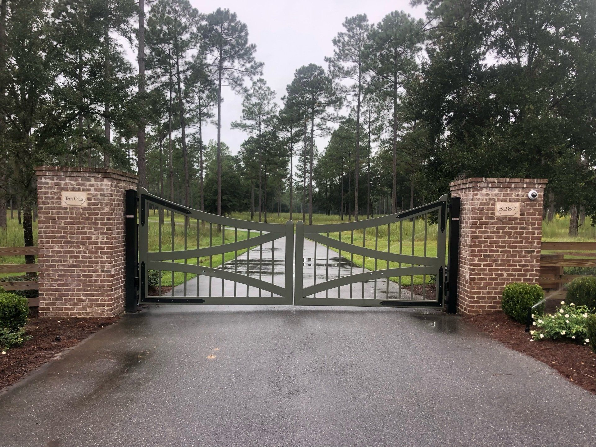 Gated entrance to a property with brick columns, black metal gates, and a paved driveway, surrounded by trees.