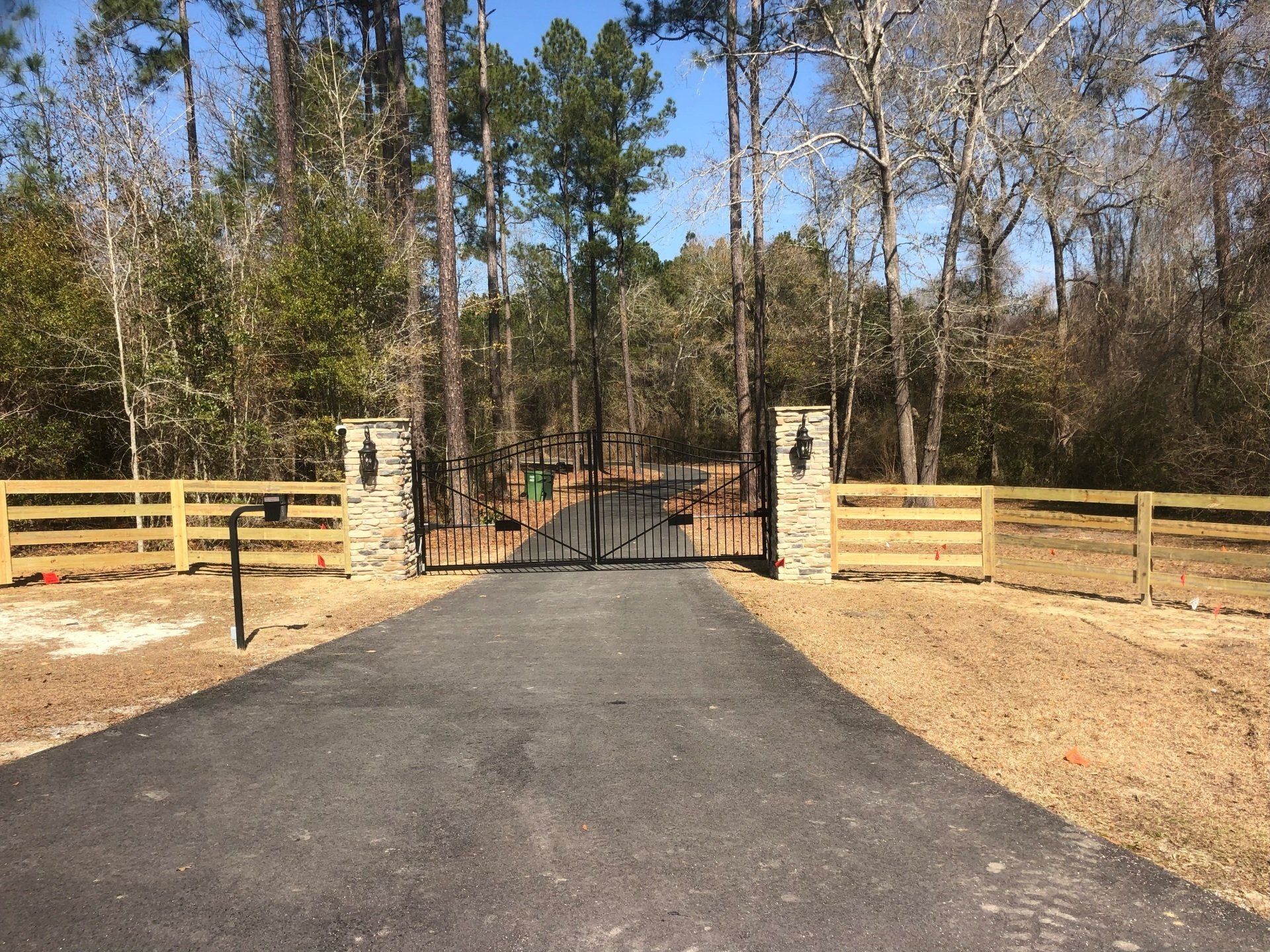 Black asphalt driveway leads to a gated entrance with stone columns and wooden fencing. Trees in the background.