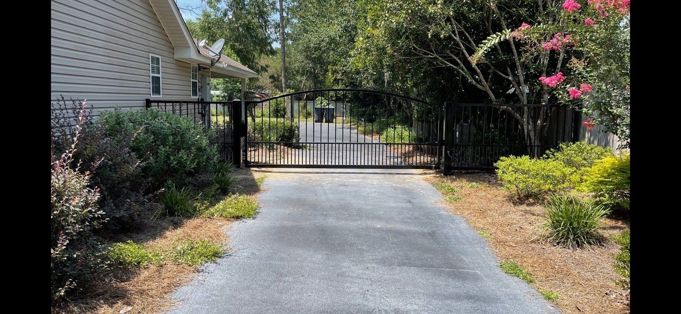 Driveway leading to black gate and house with bushes and trees.
