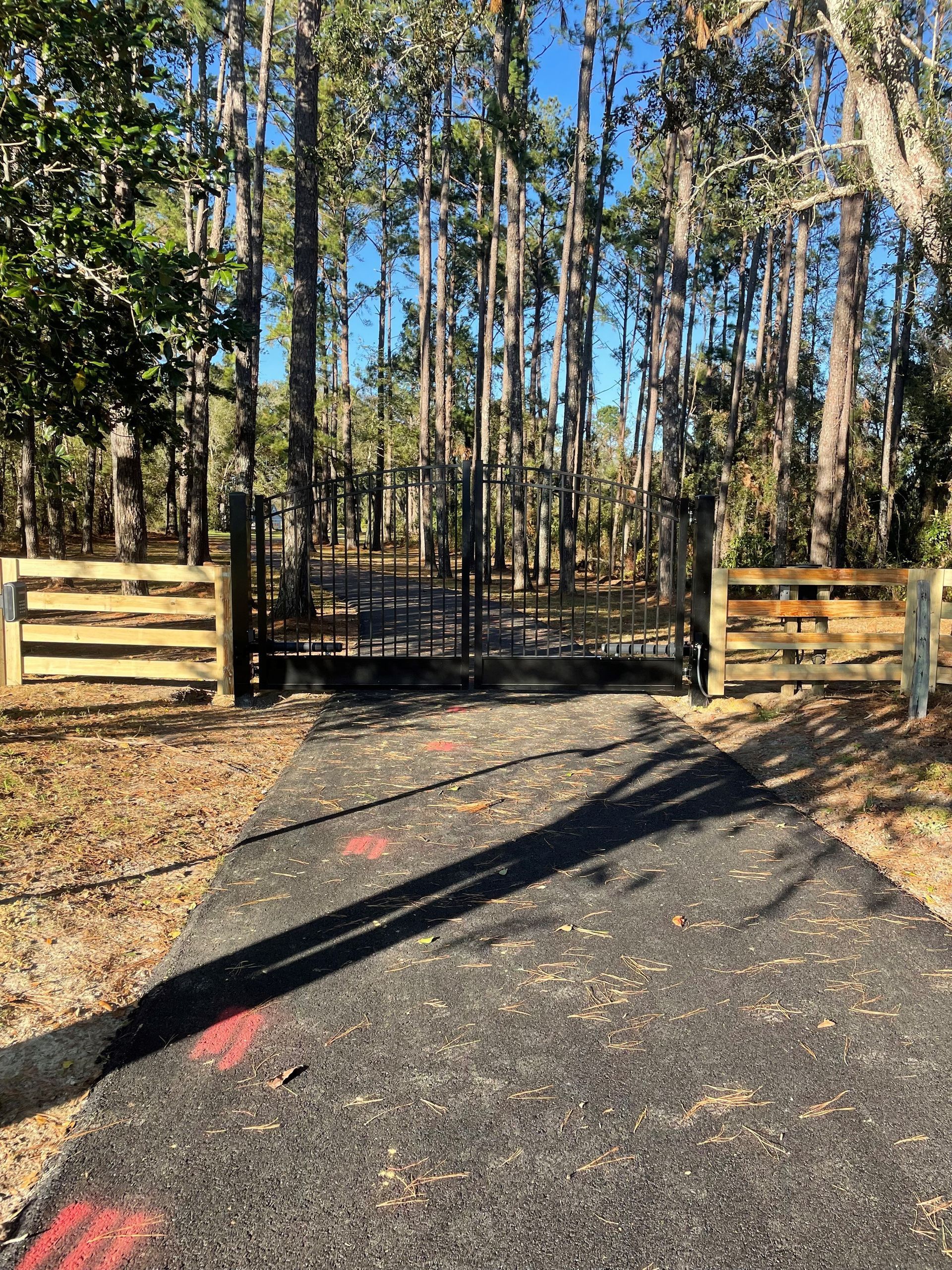 Black gate on paved driveway, flanked by wooden fences, leading into a wooded area under a bright sky.