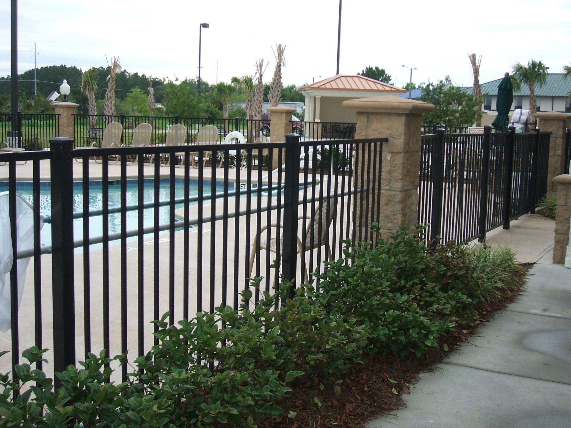 Black metal fence surrounds a pool, with decorative stone pillars. Green bushes line the fence.