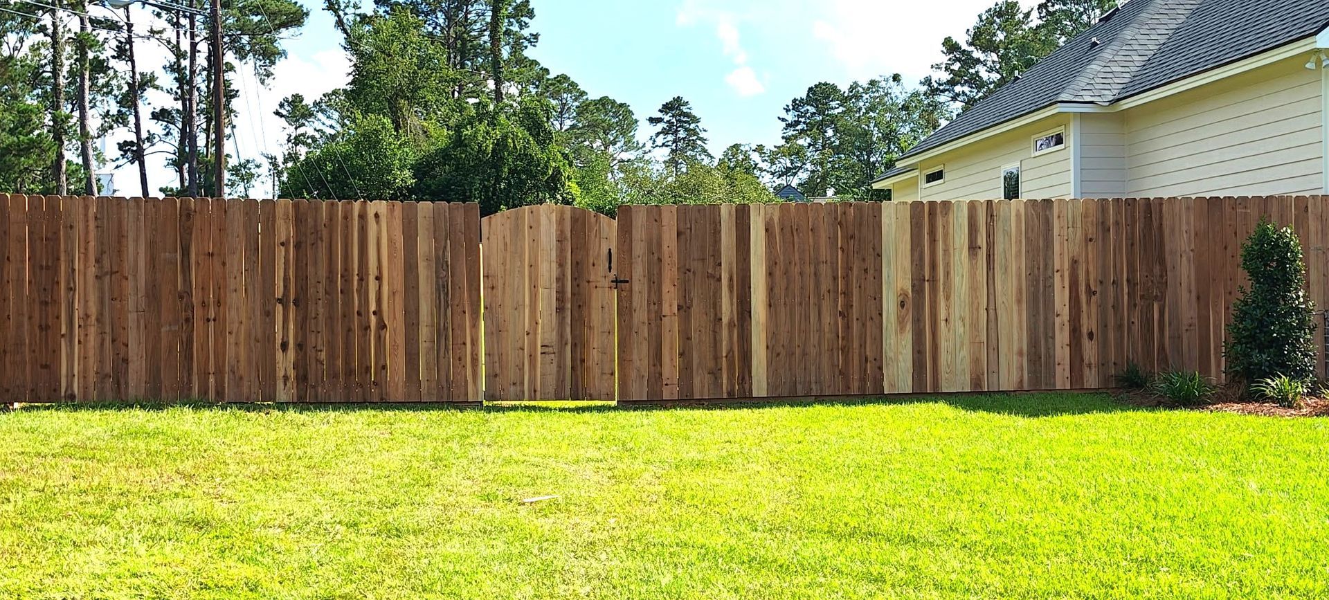Wooden fence in a backyard with a grassy lawn, trees in the background, and part of a house.