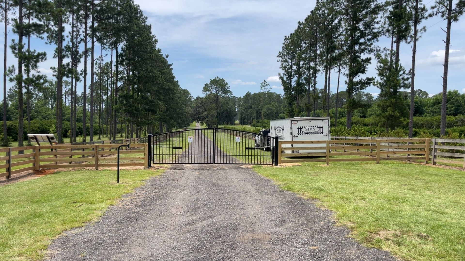 Gravel driveway leads to a gated entrance with wooden fences and trees lining the path on a sunny day.