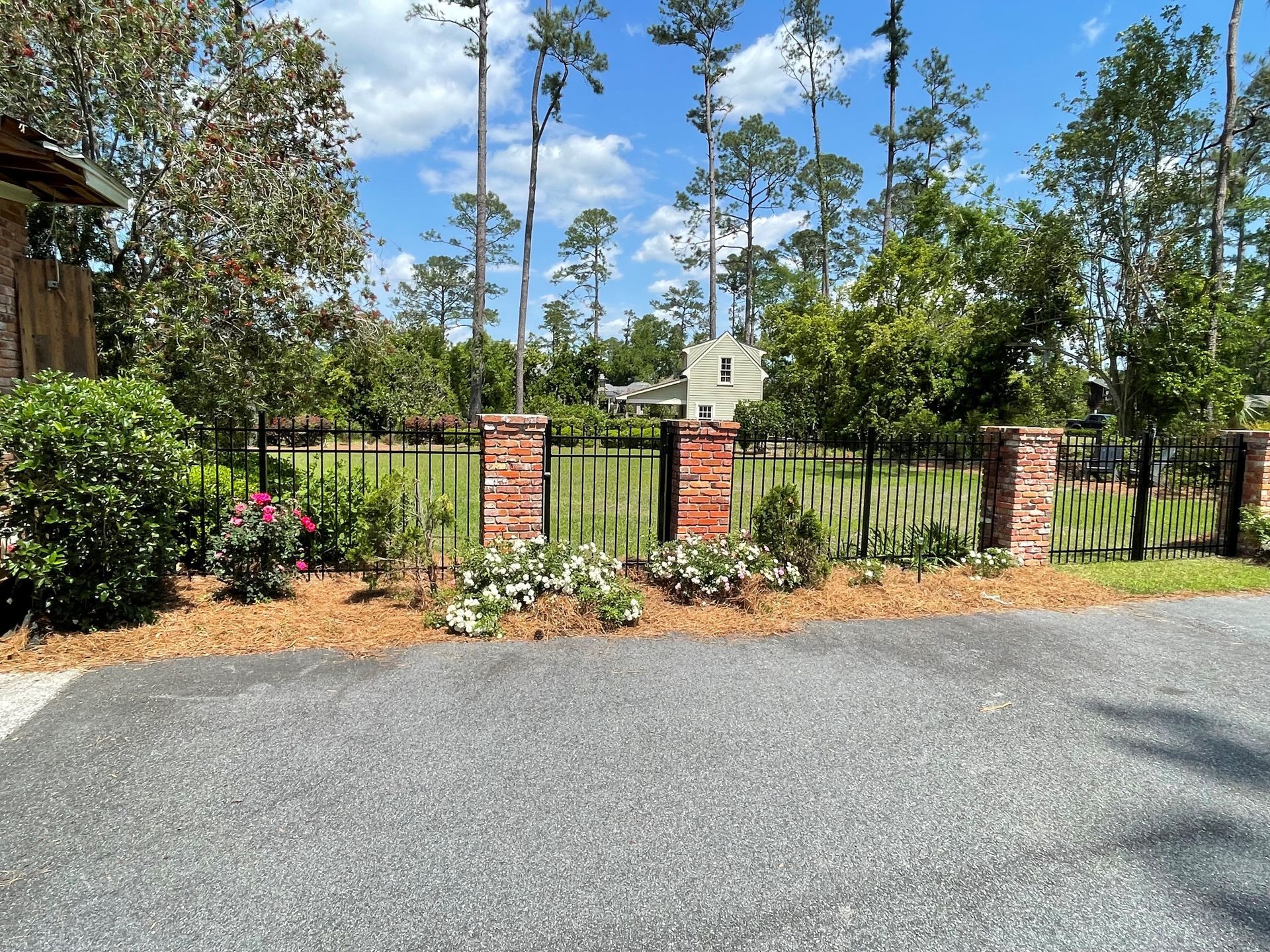 Brick pillars frame a black metal fence with landscaping, a gravel driveway, and a house in the background.