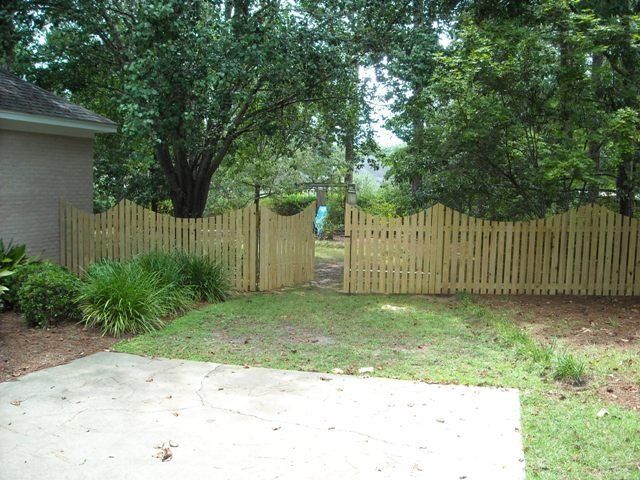 Wooden picket fence with wavy top, gate open, leading to wooded area. Concrete patio in foreground.