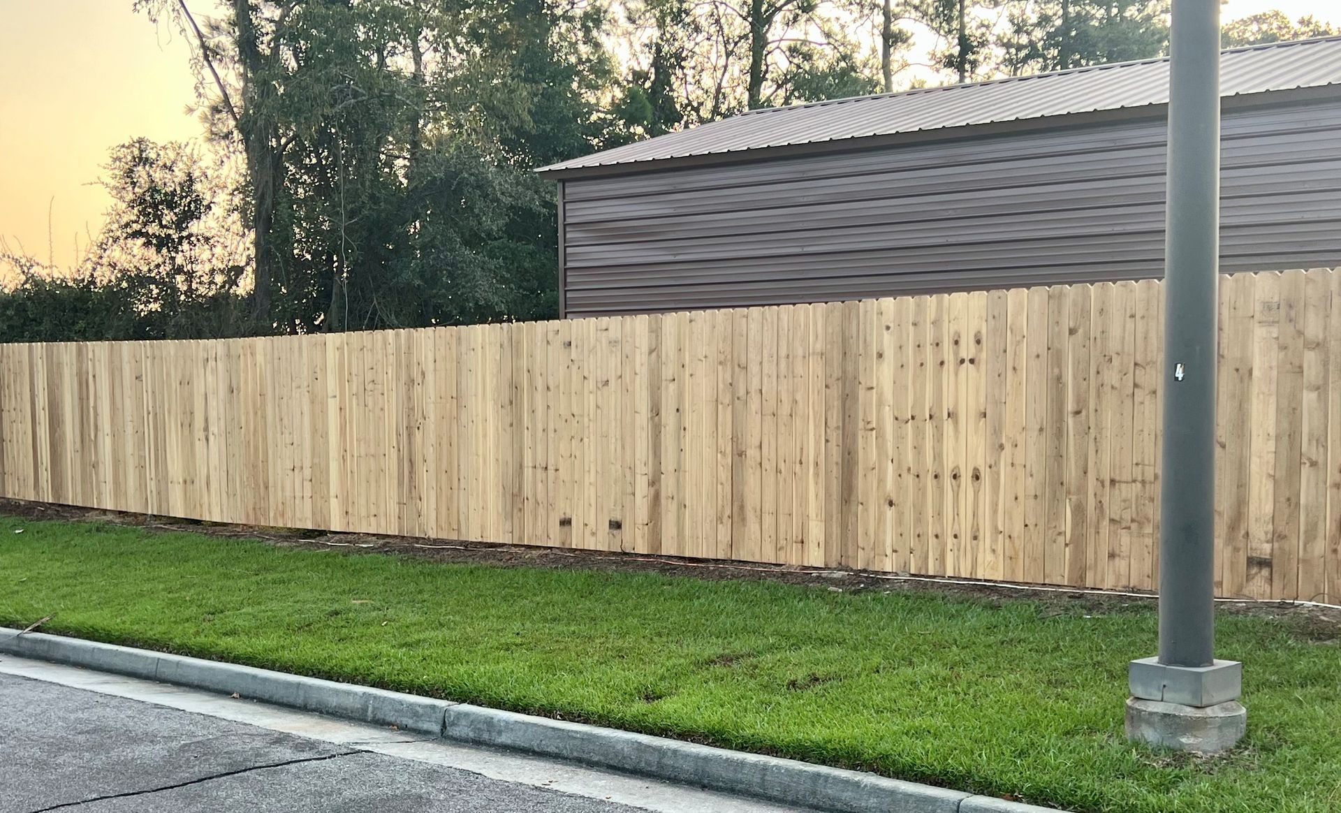 Wooden fence bordering green grass with a brown building in the background and a street in front.