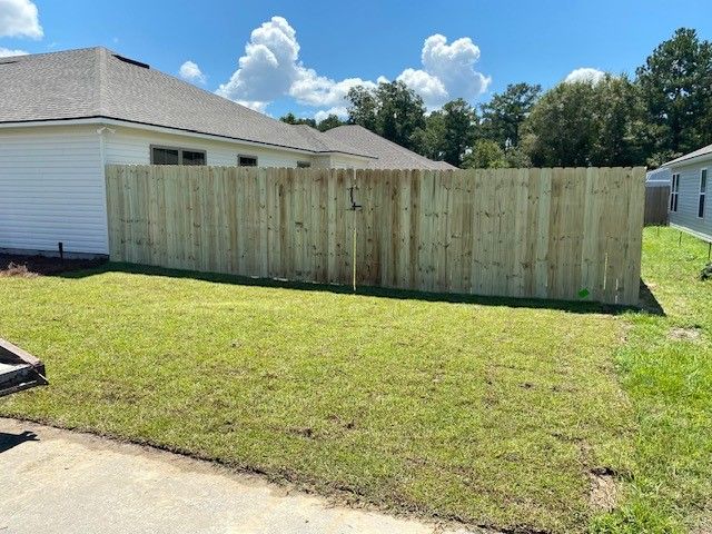 Wooden fence in a backyard with green grass and a house. Blue sky with clouds in the background.