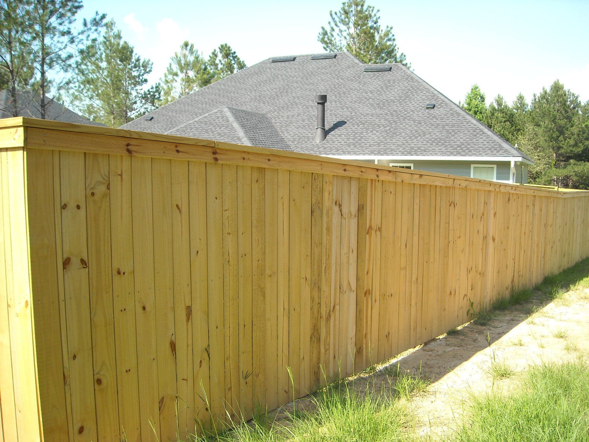 Wooden fence surrounds a house with a gray roof and trees in the background.