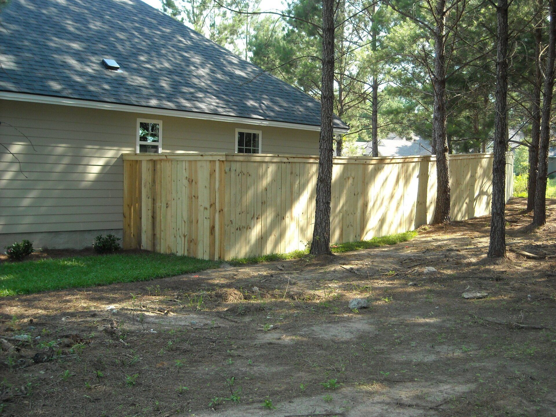 Wooden fence bordering a beige house, next to trees and grass.