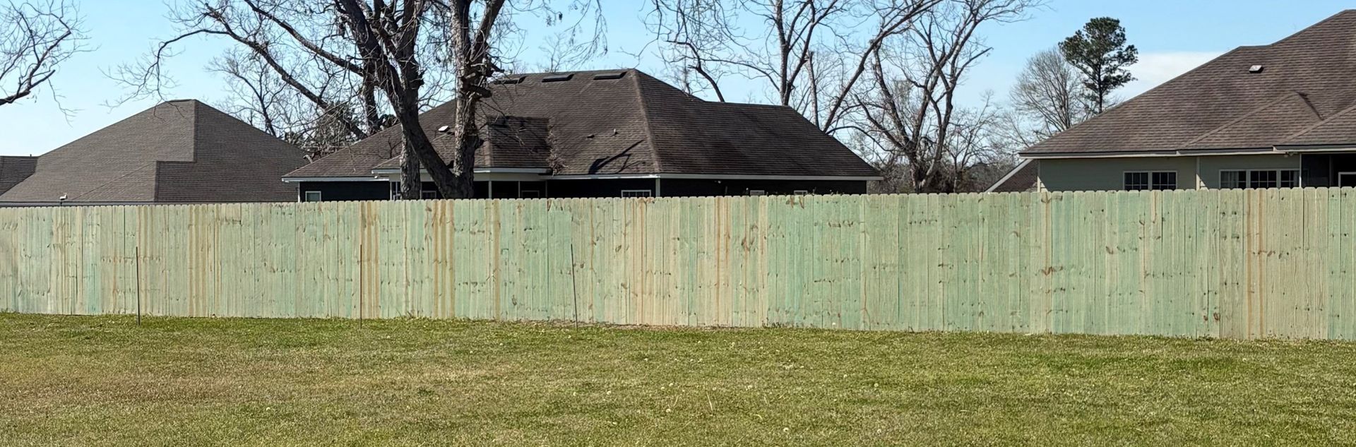 Wooden fence in front of grass with houses and bare trees in the background. Bright blue sky.