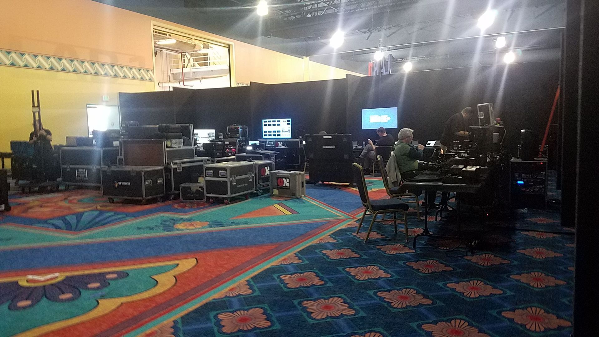 A production room with equipment and crew; colorful patterned carpet, black screens, stage lights, and boxes.