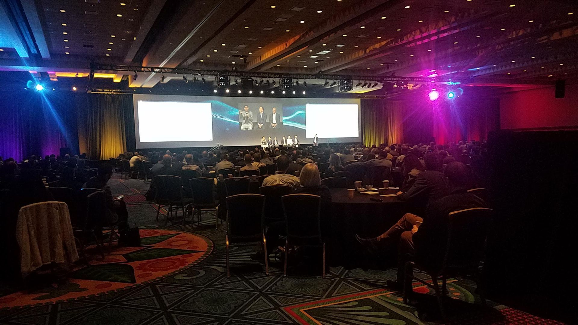 Large event hall with stage and audience. People in seats, screen displays, colorful lighting.