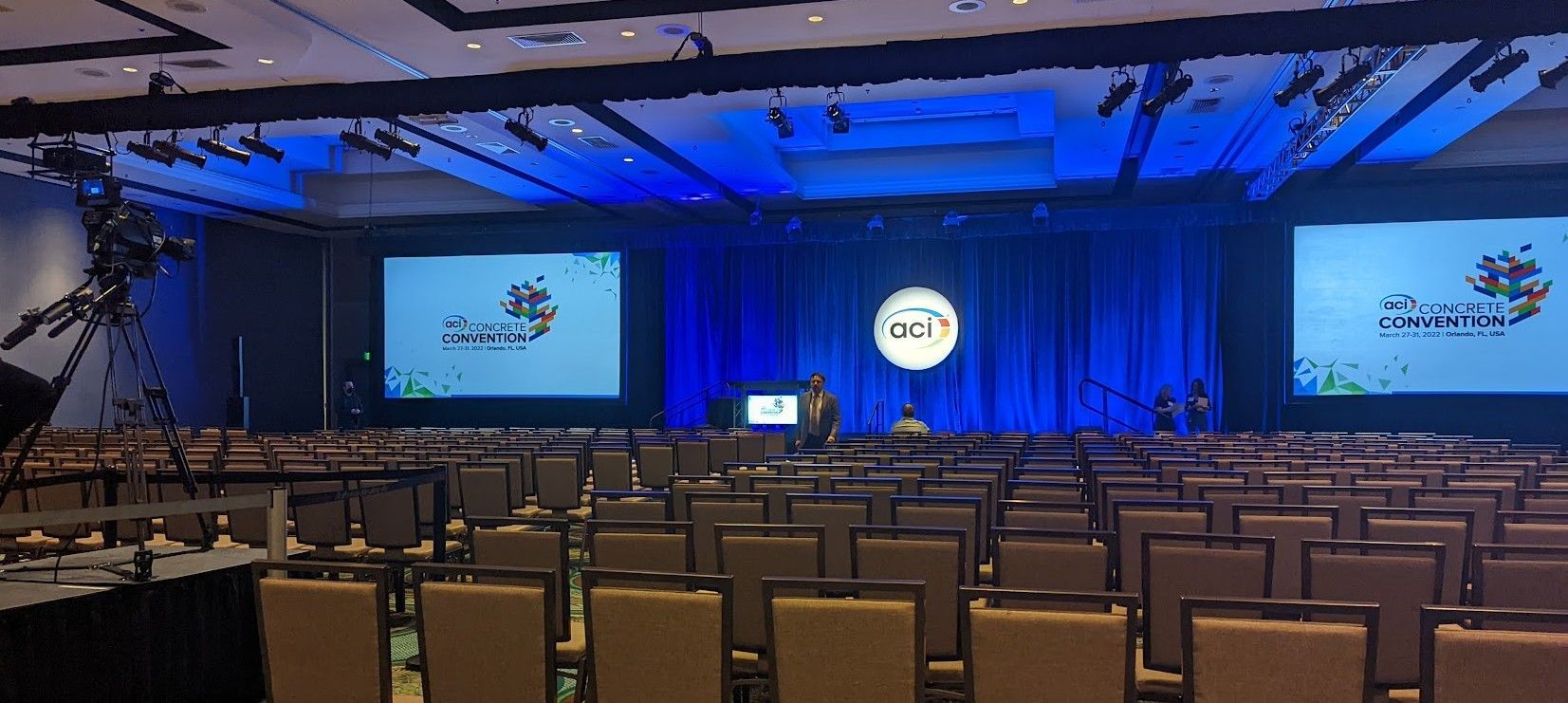 Conference hall with rows of seats facing a stage with two screens and a presenter. Blue lighting.