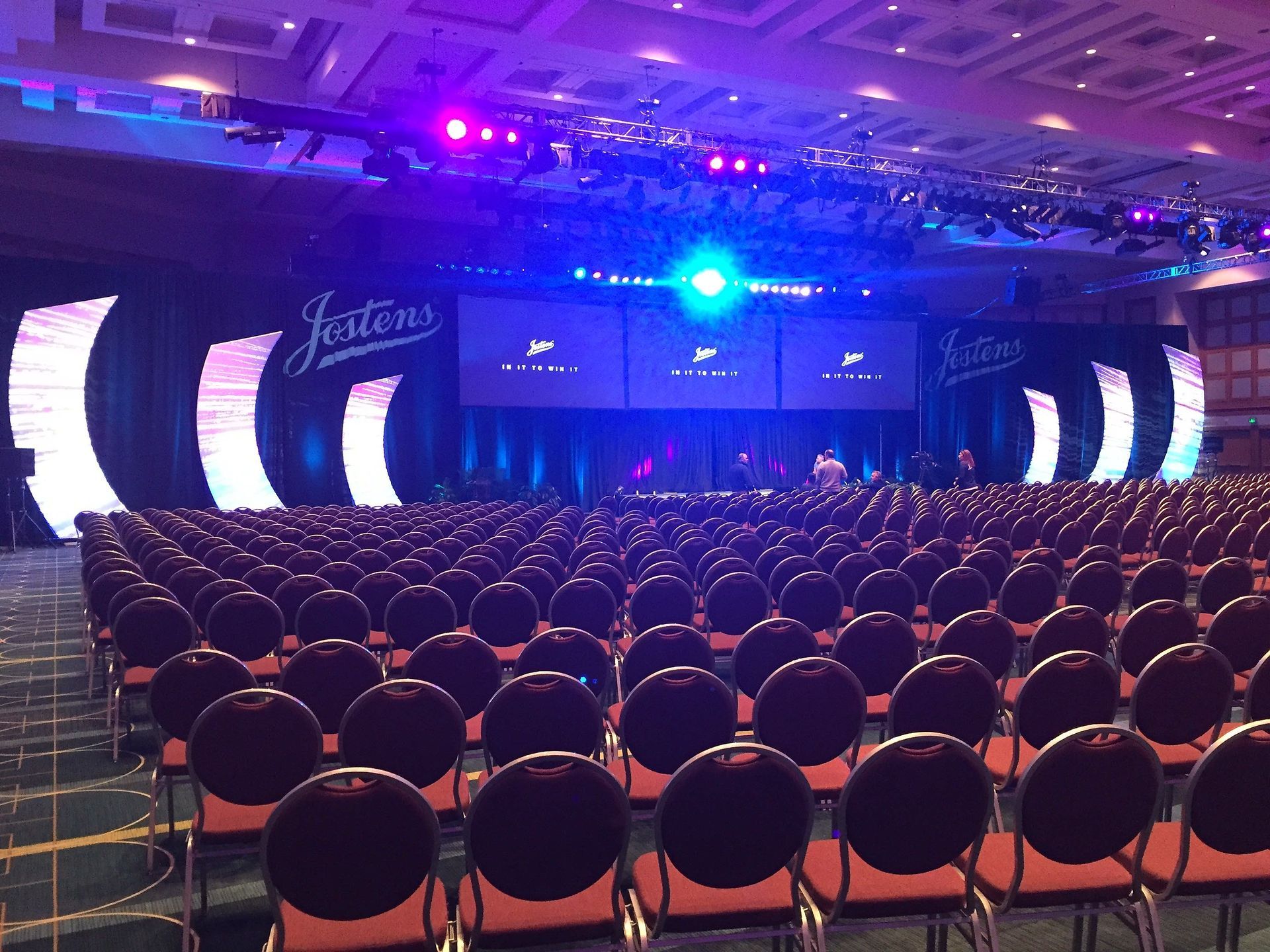An empty convention hall set up for a presentation, with rows of chairs facing a stage with screens and lighting.