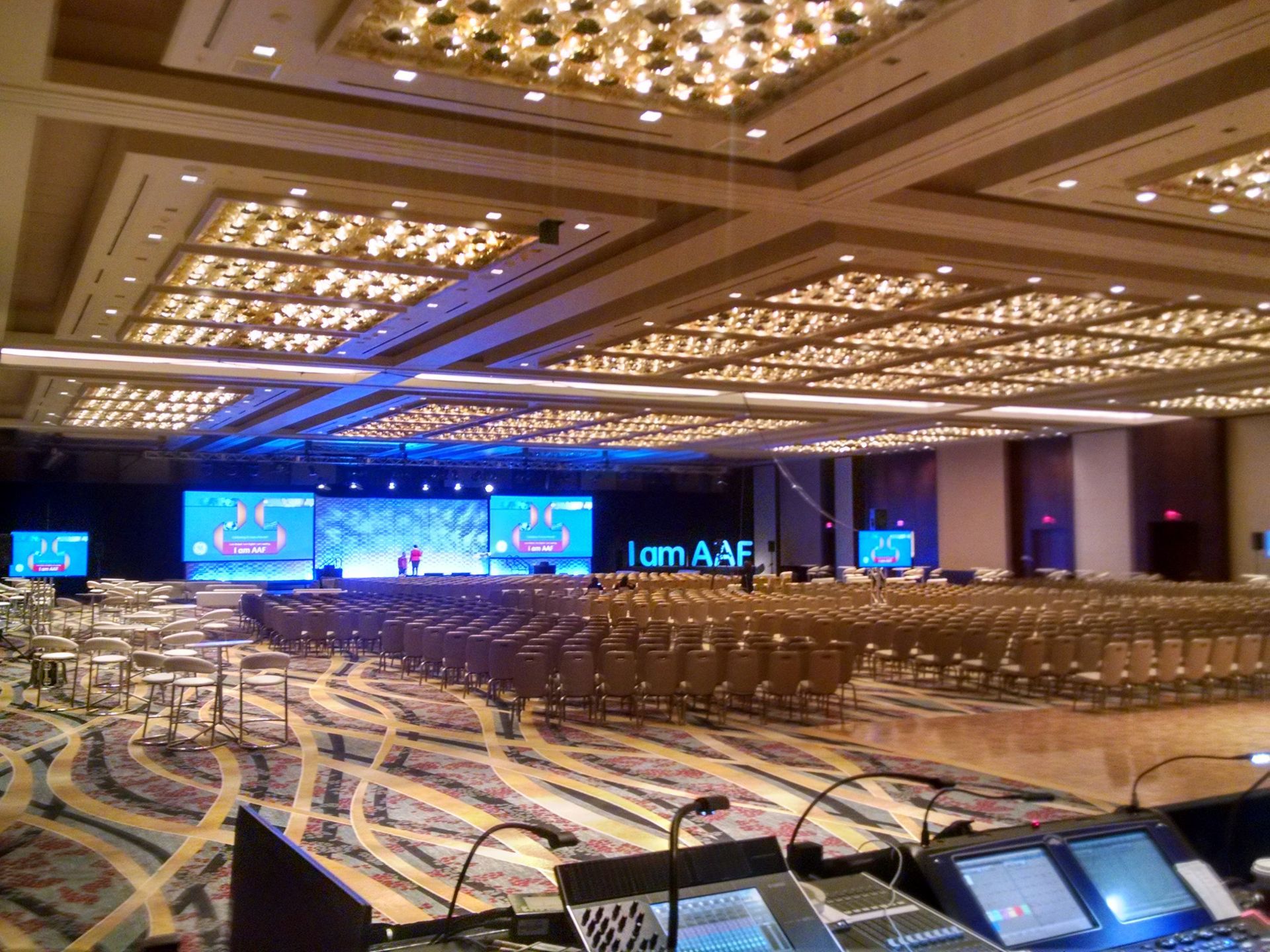 Large conference hall with a stage set for a presentation; rows of chairs face the stage, soundboard in foreground.