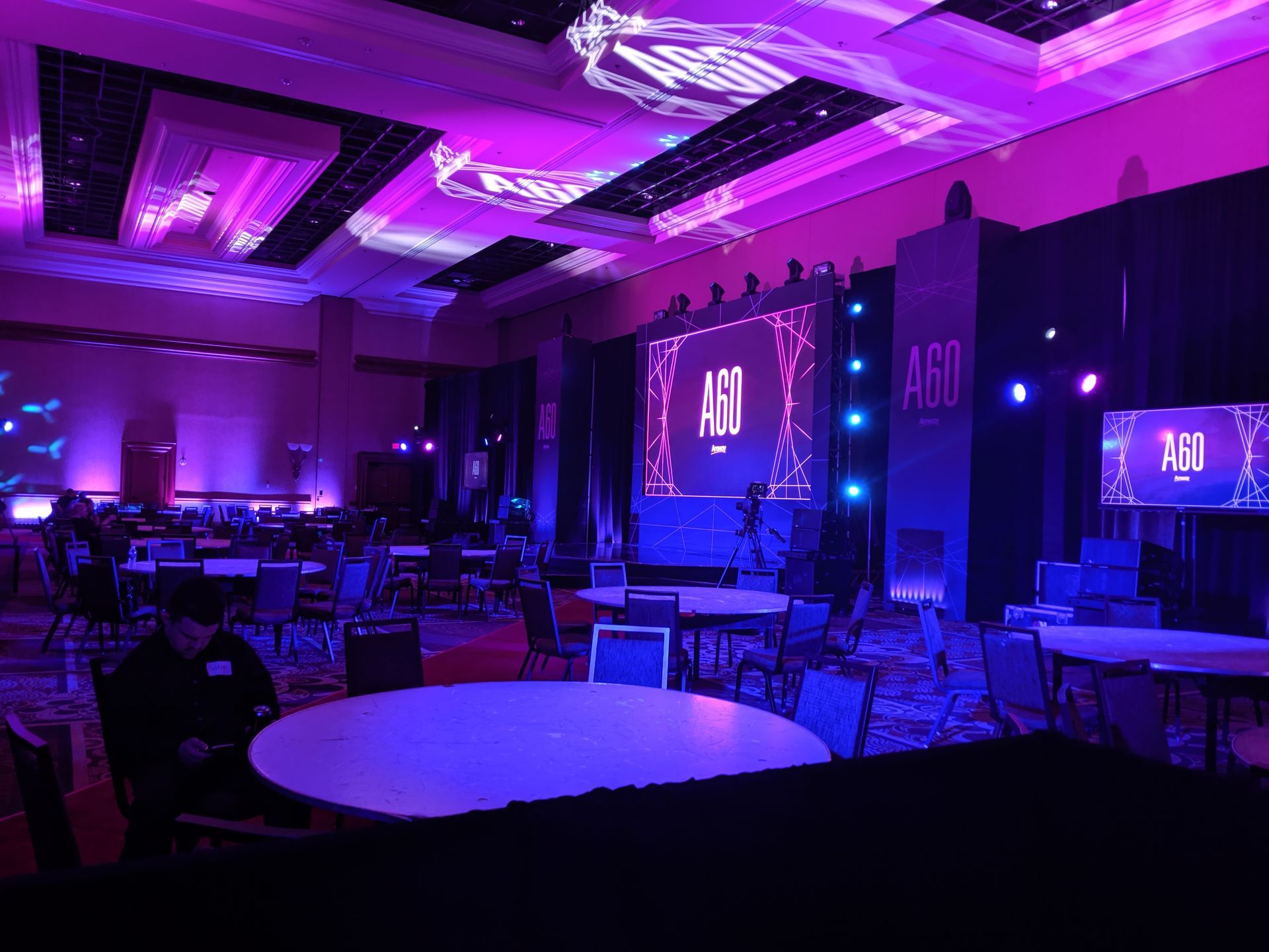 A large ballroom with round tables, a stage with purple lighting, and the text 