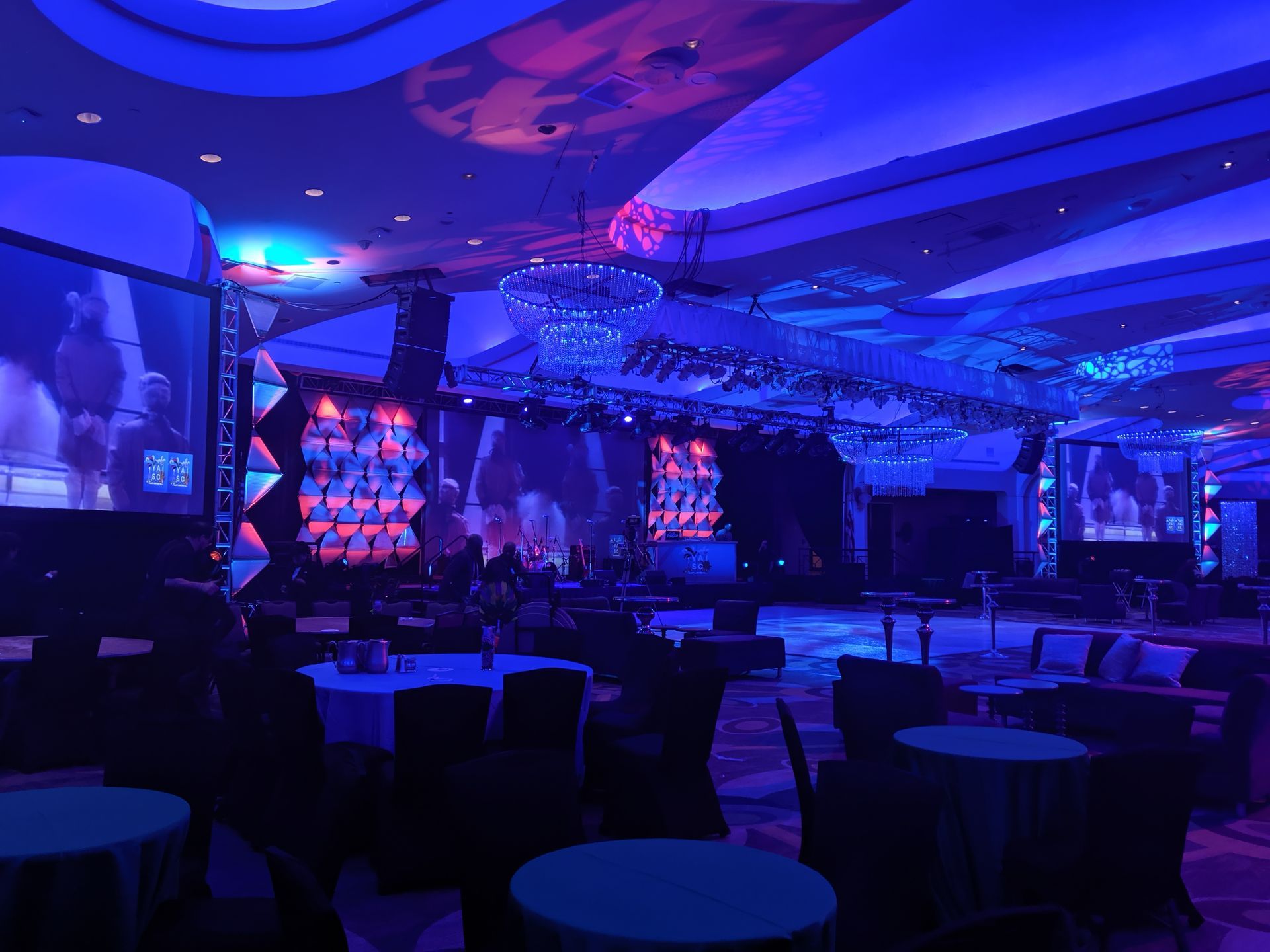 Blue-lit event hall with tables, stage, screens, and chandeliers.