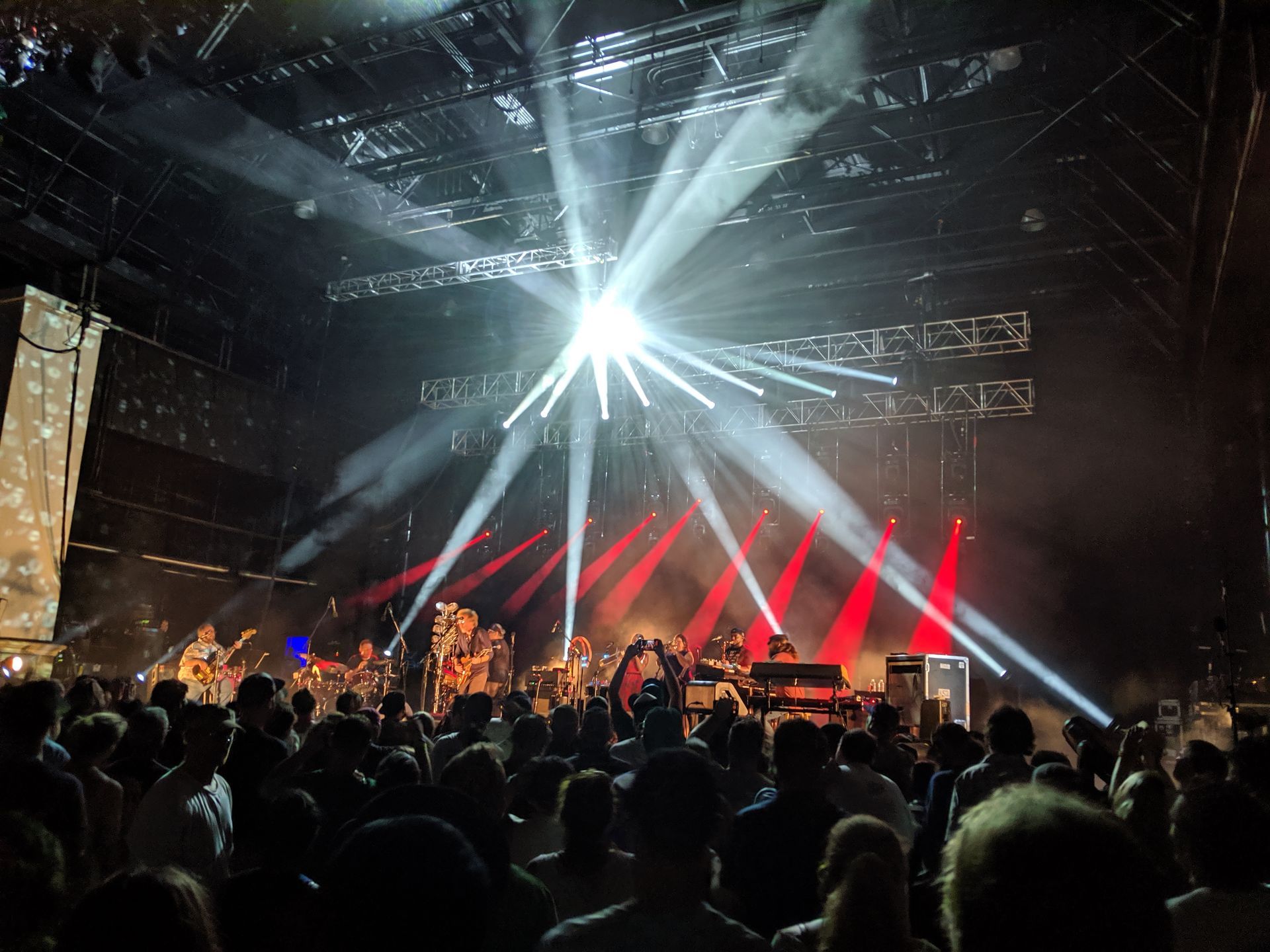 Concert stage with band performing under bright stage lights, viewed from a crowd of people.