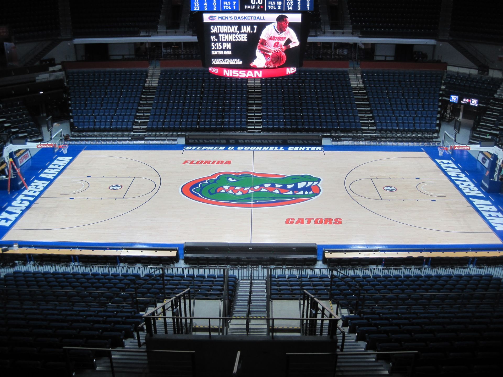 Empty basketball arena, center court with a large Florida Gators logo. Blue seating and scoreboard overhead.