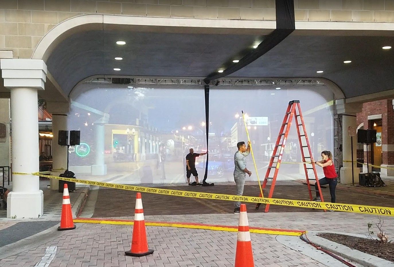 People setting up an outdoor projection screen under an arched structure, using a ladder. Road closed with cones and tape.