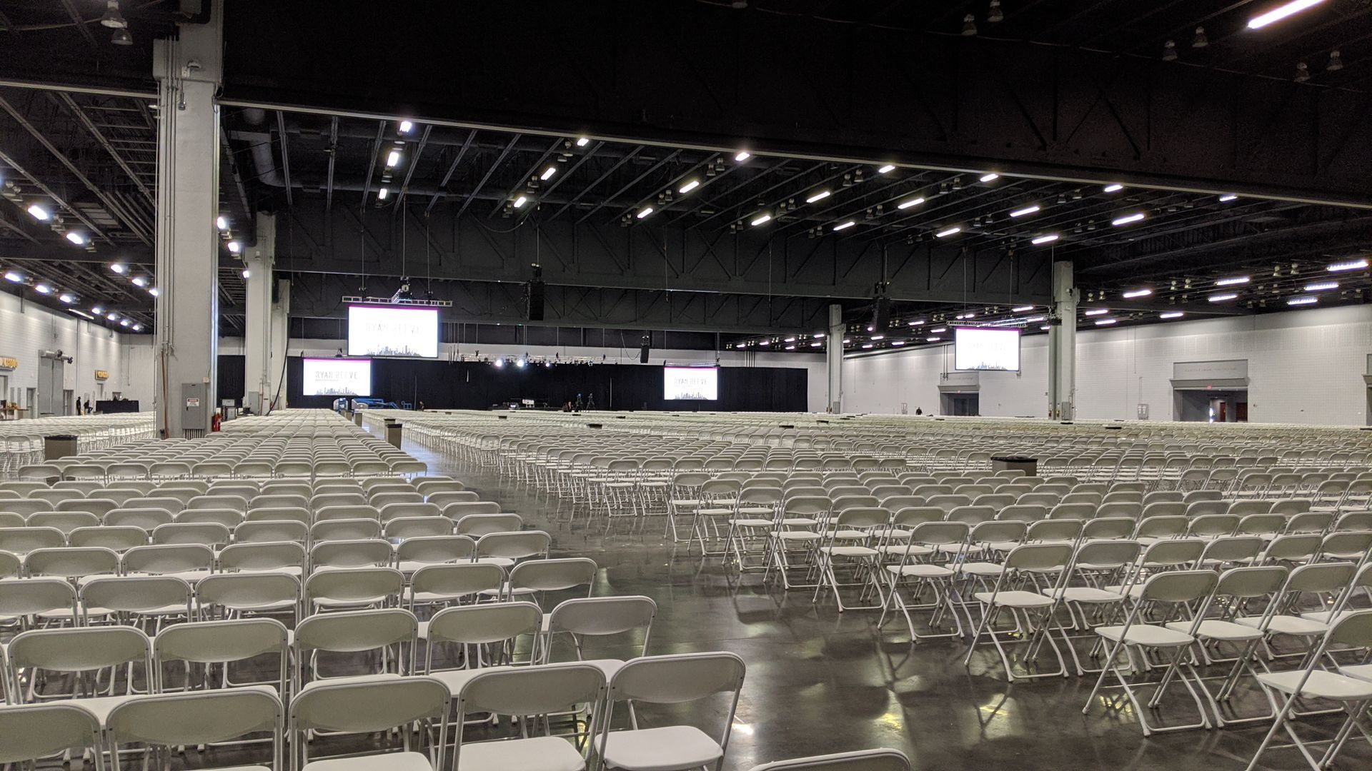 Rows of empty chairs fill a large convention hall, with screens on the back wall, ready for an event.