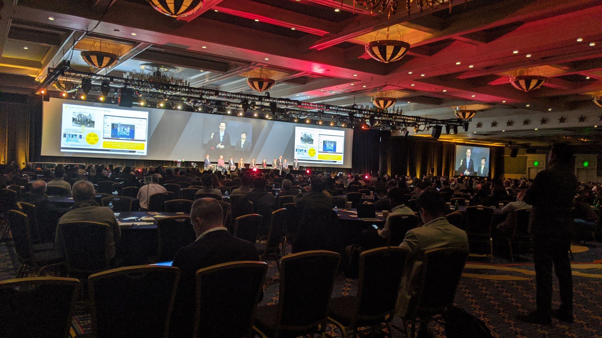 Conference hall with presenter on stage, large screen, and seated audience.