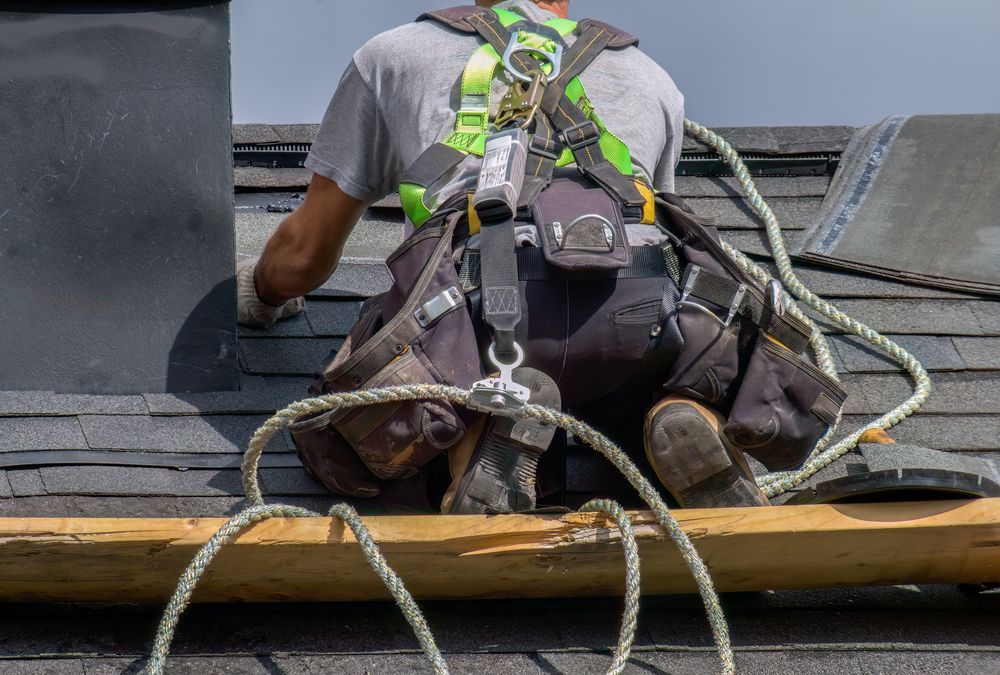 Un operaio edile, con indosso un'imbracatura di sicurezza e una cintura porta attrezzi, lavora su un tetto di tegole accanto a un camino.