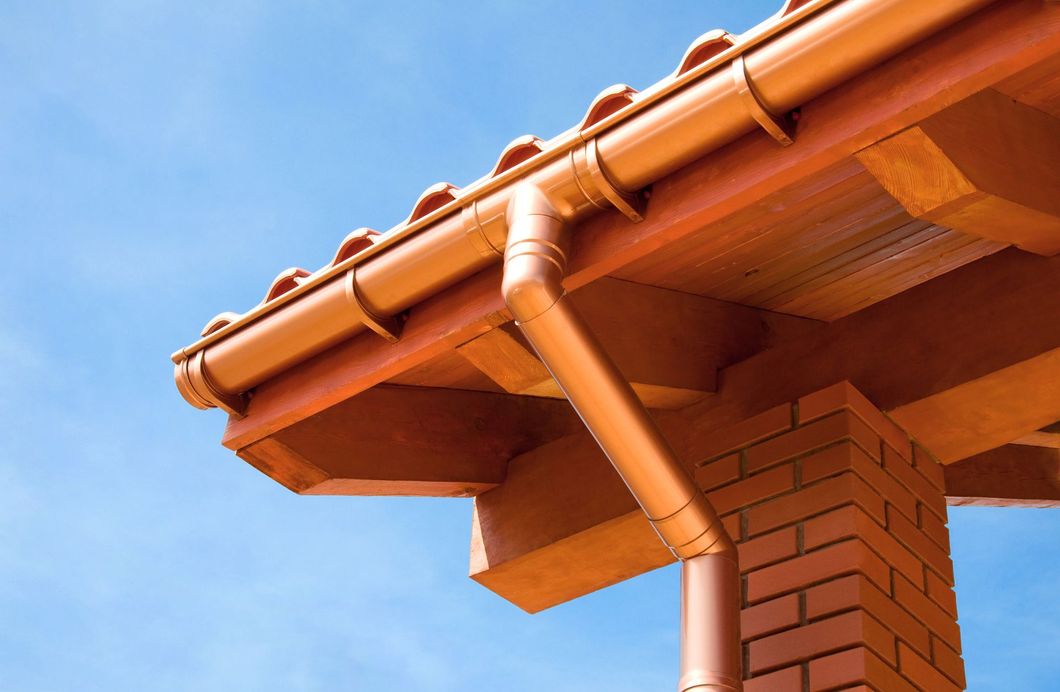 Sistema di grondaie in rame installato sul bordo di un tetto di tegole in terracotta e su un pilastro in mattoni, contro un cielo azzurro limpido.