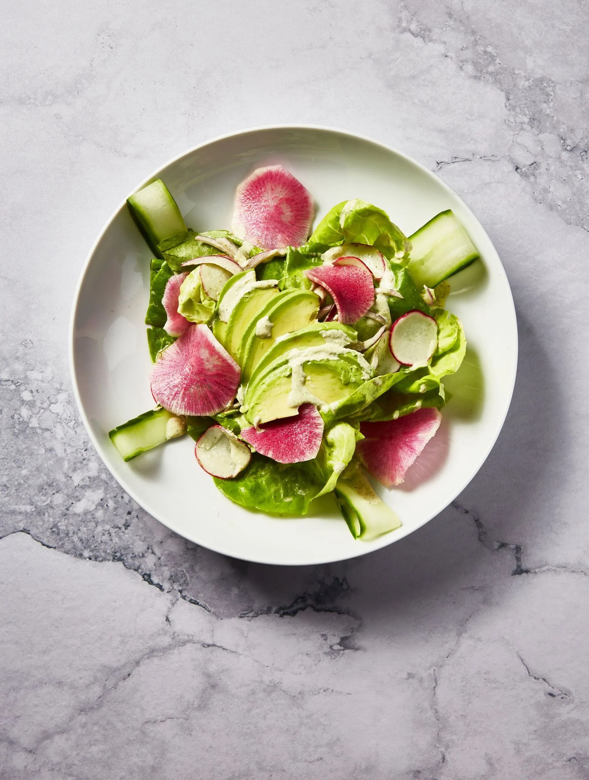 Avocado, watermelon radish, cucumber, and lettuce salad in a white bowl on a marble surface.