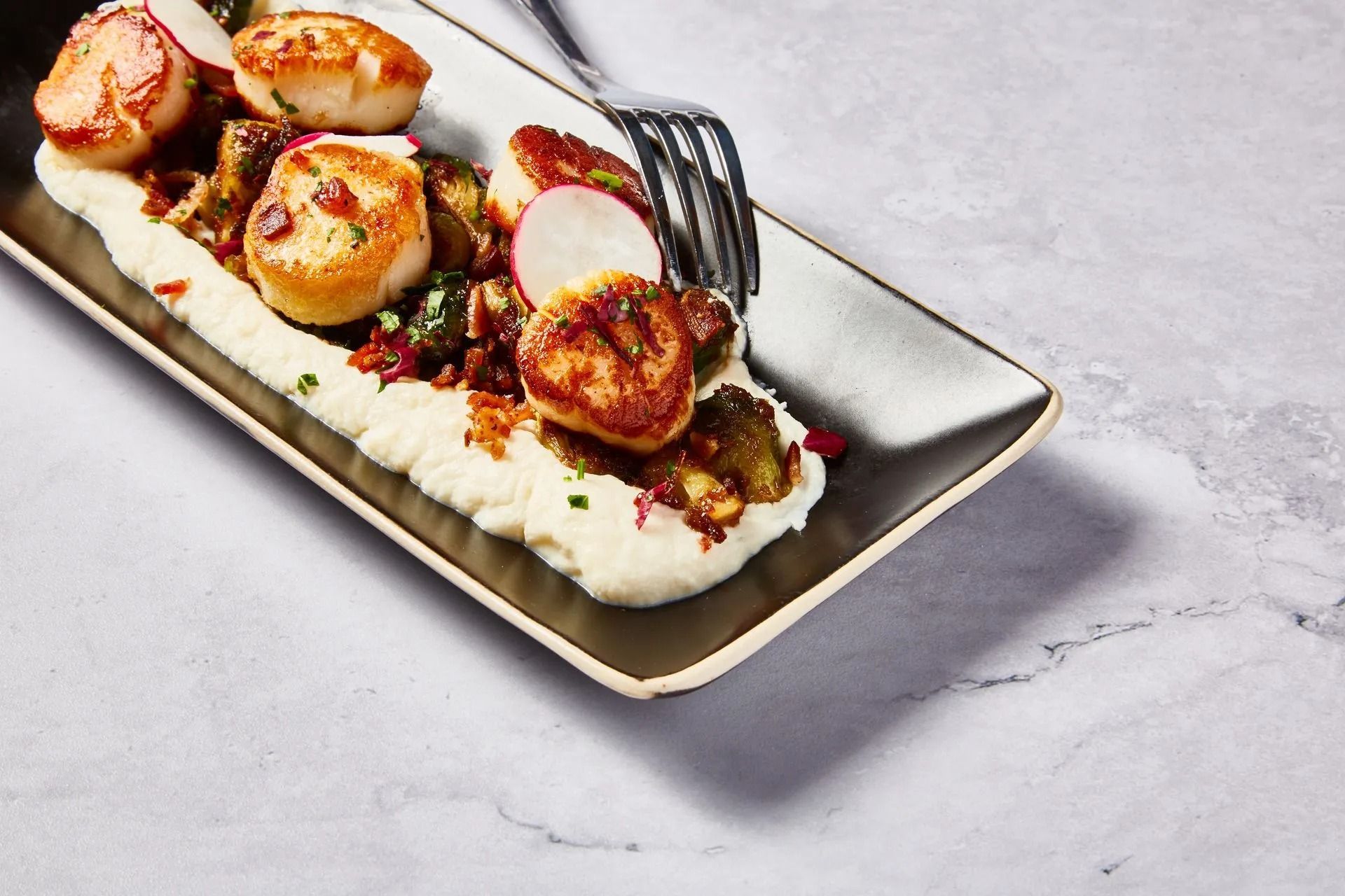 Seared scallops atop mashed potatoes, Brussels sprouts, and radish slices, served on a rectangular plate.